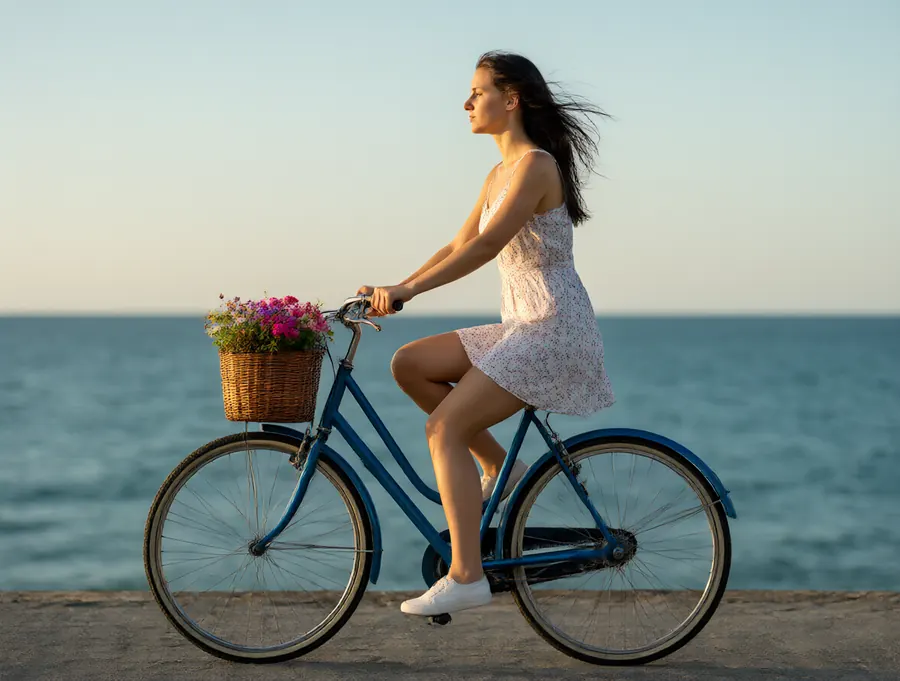 A young woman on a blue bicycle with a flower basket by the sea at golden hour